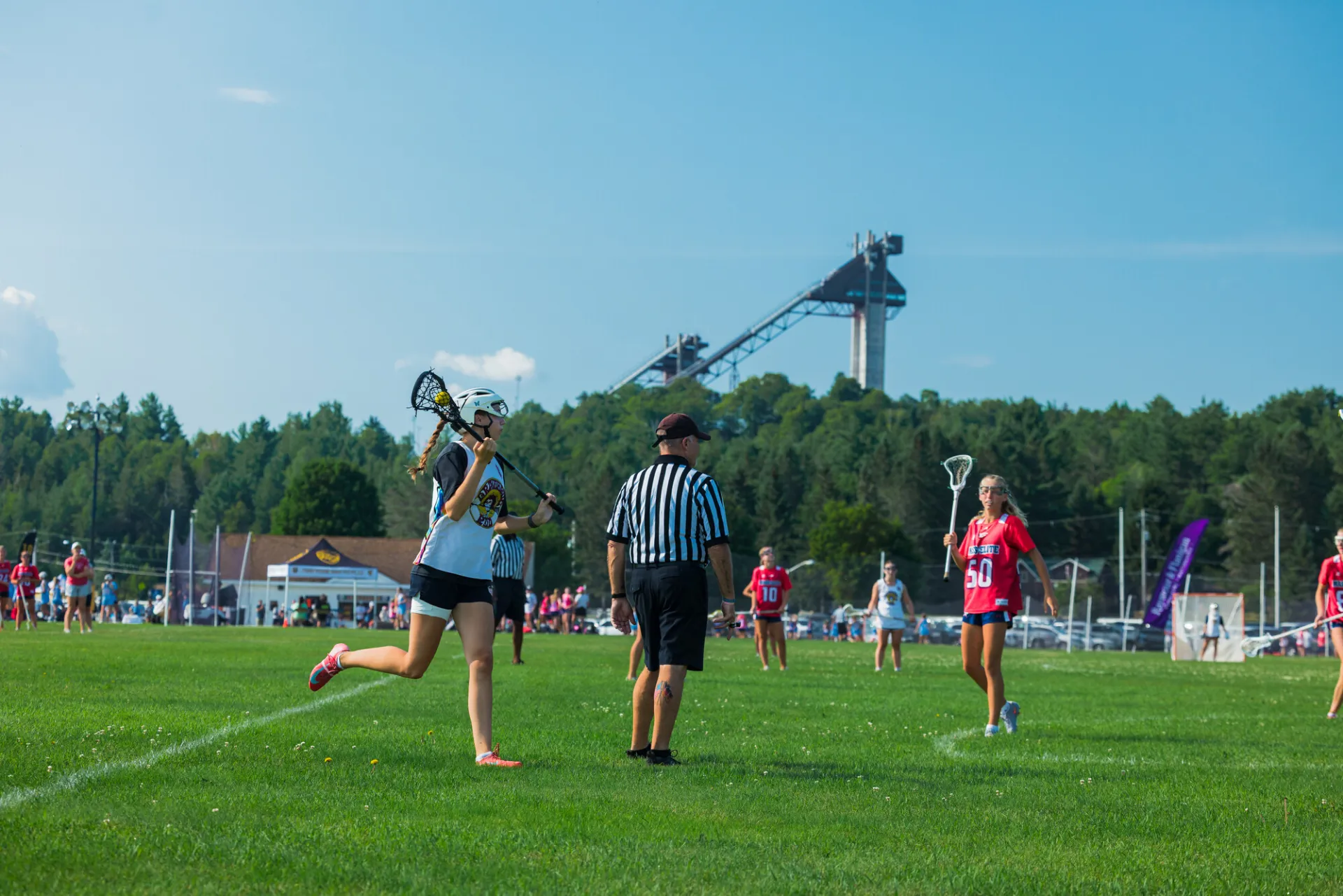 Woman in white and black uniform holds lacrosse ball in stick while running past referee during game on field in Lake Placid