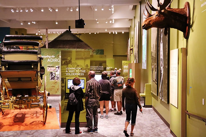 A large room with green walls where people are viewing the plaques under various displays like a carriage and a moose.