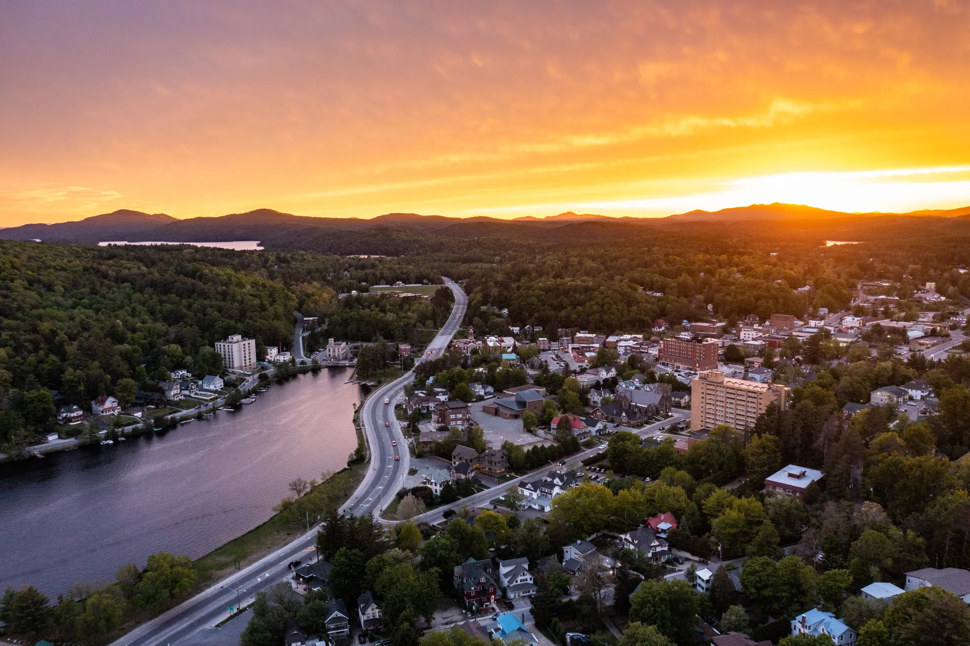An aerial image of a sunset over a small Adirondack town. A lake appears on the left next to a winding road.