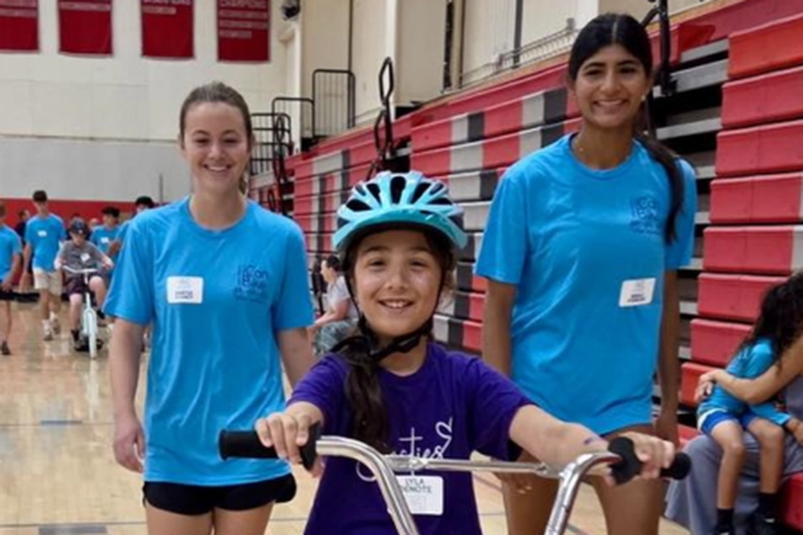 A child with blue helmet smiles on a bike in a gymnasium flanked by two volunteers.