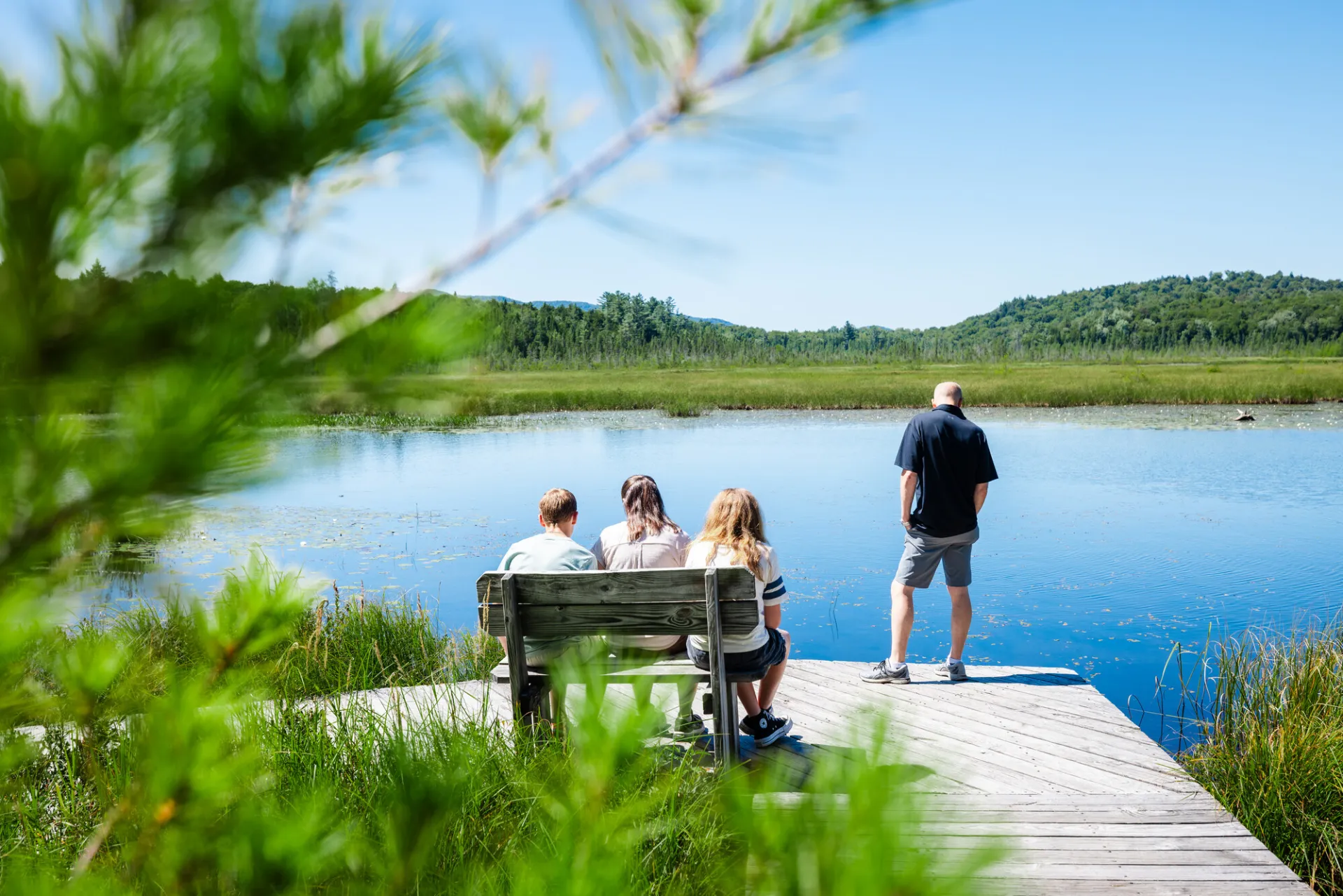 Three people sit on a bench while one person stands on a dock overlooking a calm lake.
