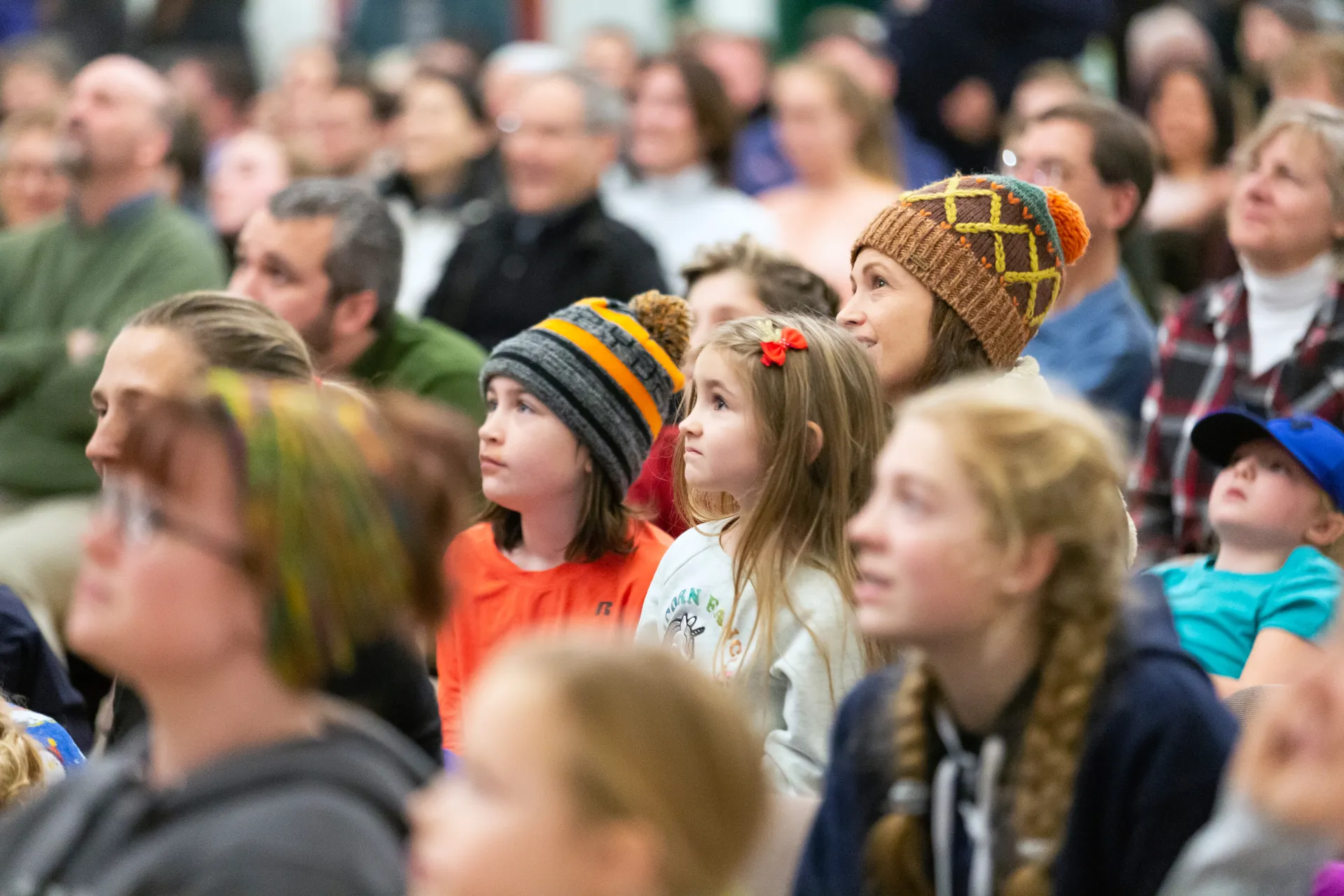A group of people, including children and adults, attentively listen in an indoor setting.