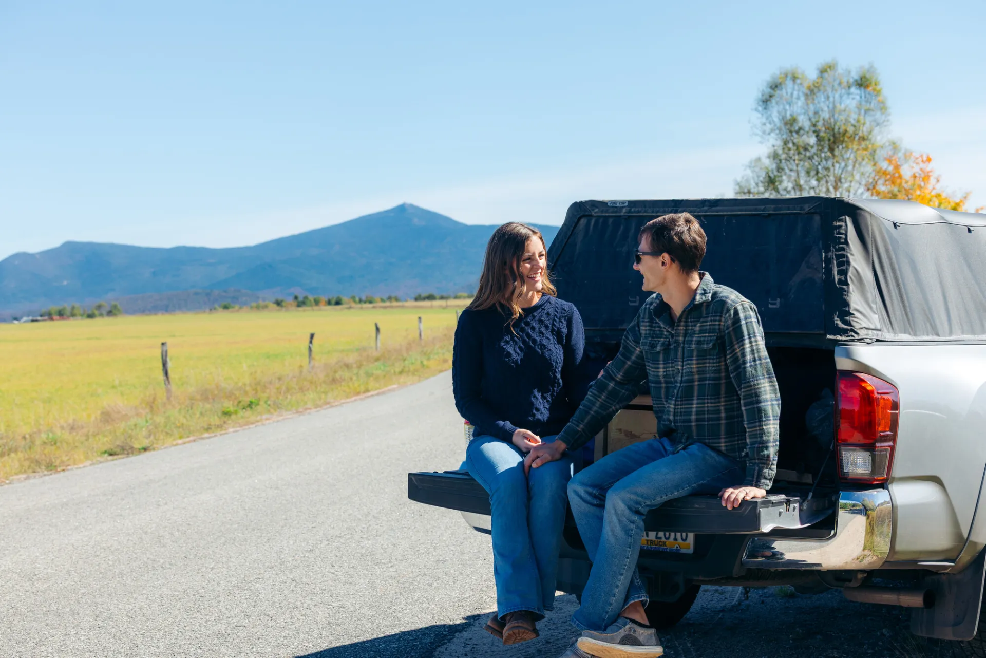 A woman and man sit on the tailgate of a truck with mountains in the background. 