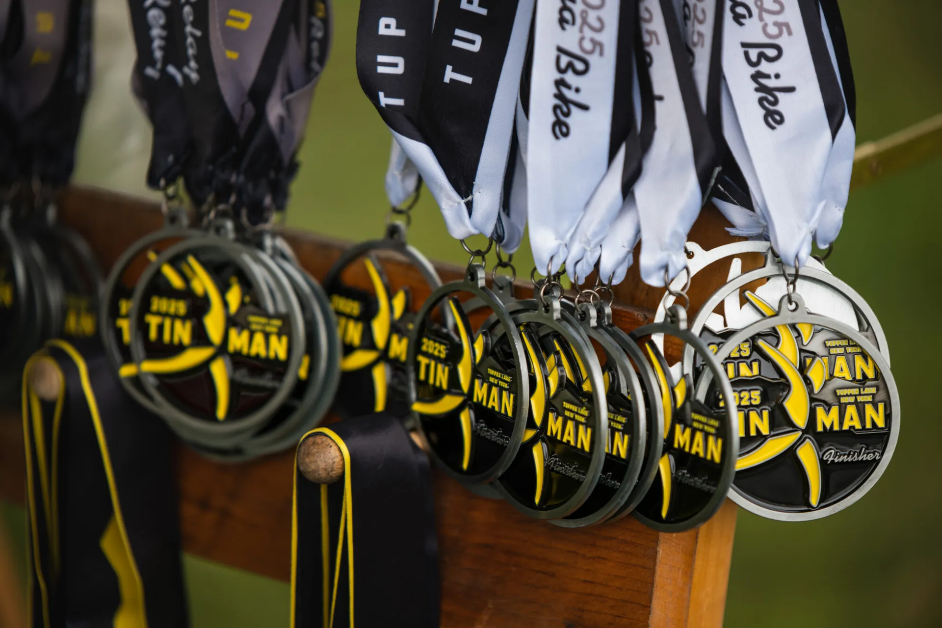 A row of silver 2025 Tupper Lake Tinman finisher medals with yellow and black accents hang from ribbons on a wooden rack.