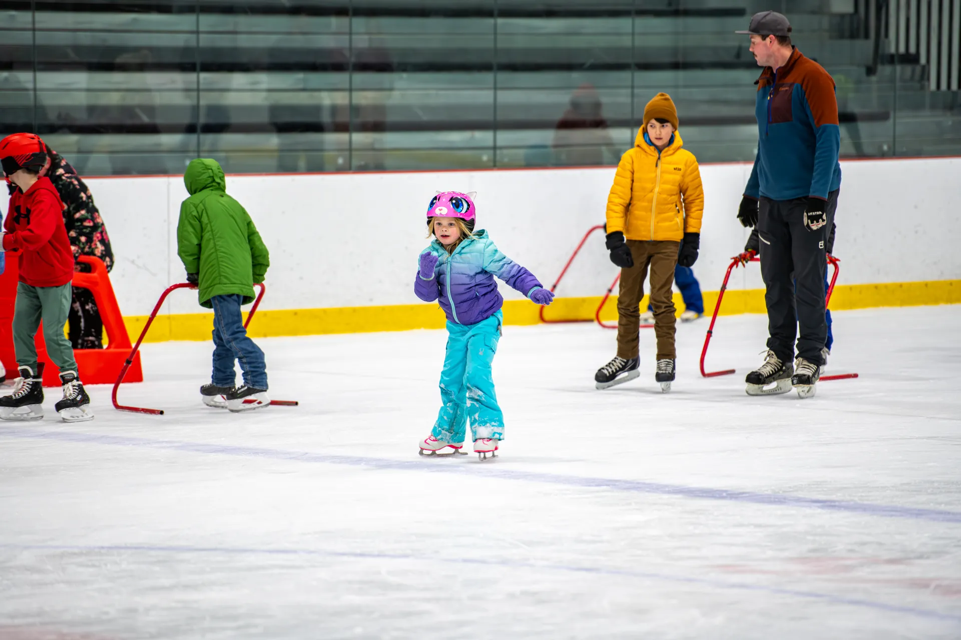 A child in a bright outfit skates without training bar while other still use them in the background.