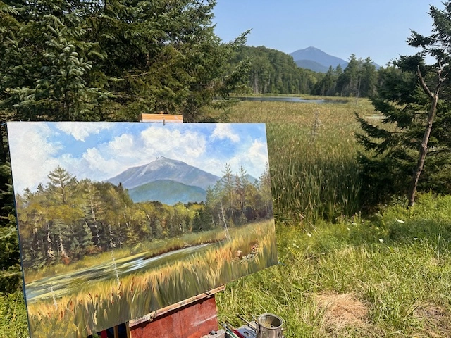 The artist's view of a plein air painting underway while still attached to an easel while still overlooking the field, pine trees, swamp surrounding a river and mountain view that inspired the art.