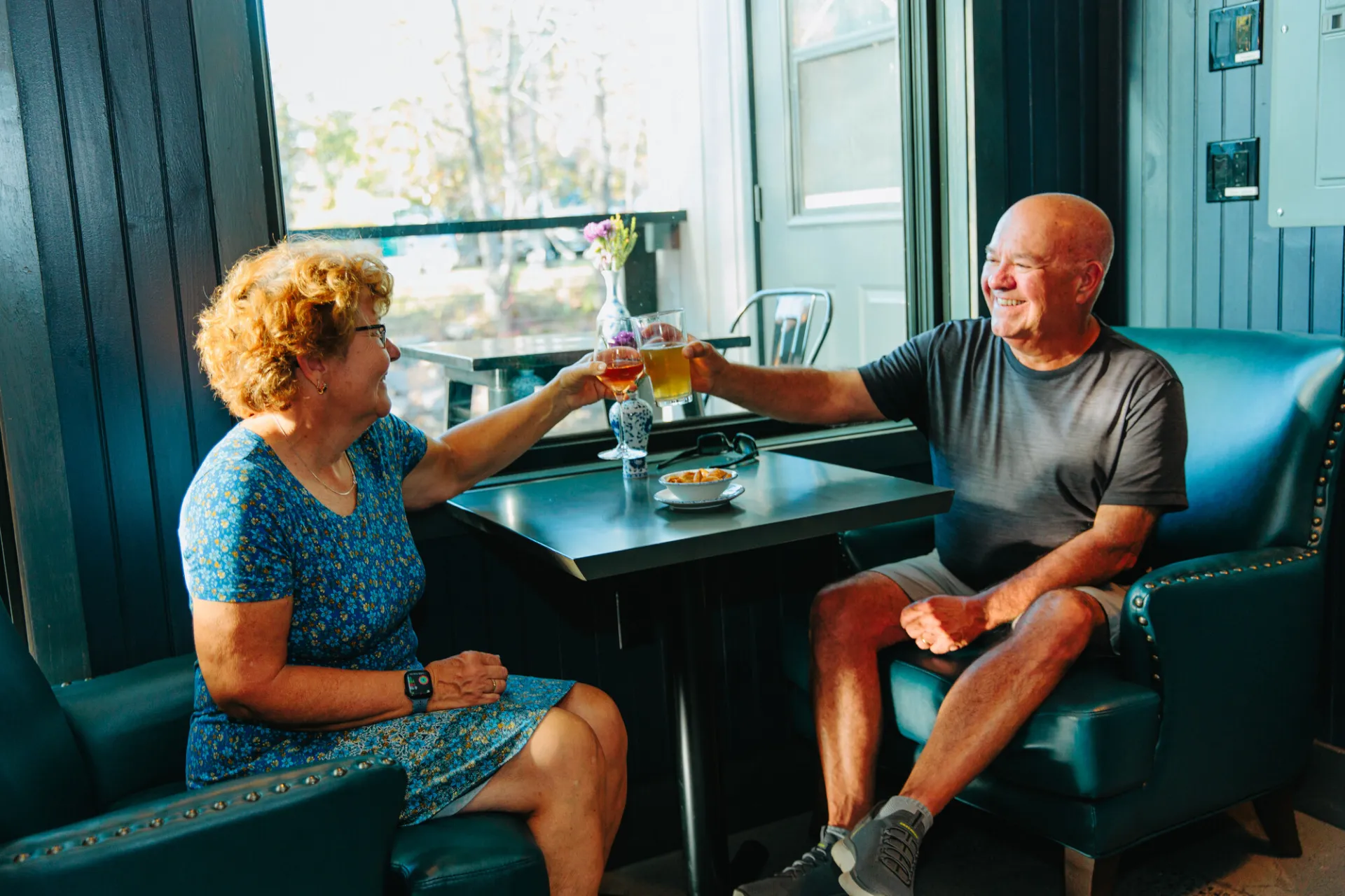 A smiling man and woman sit in plush leather armchairs by a window, clinking a wine glass and a beer glass over a table.