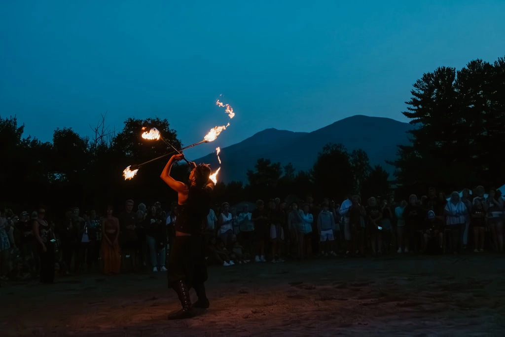 Fire dancer seen performing against backdrop of mountains and music festival at dusk