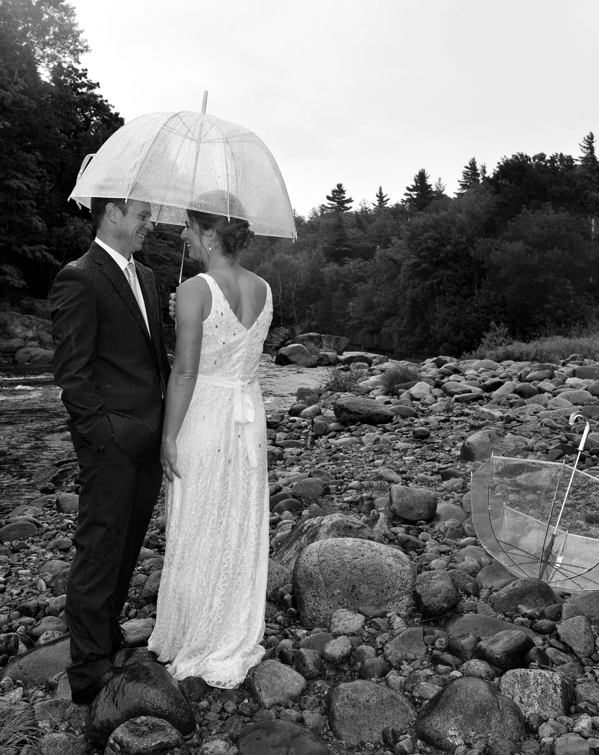 A bride and groom stand by a riverbed under a clear umbrella in black and white.