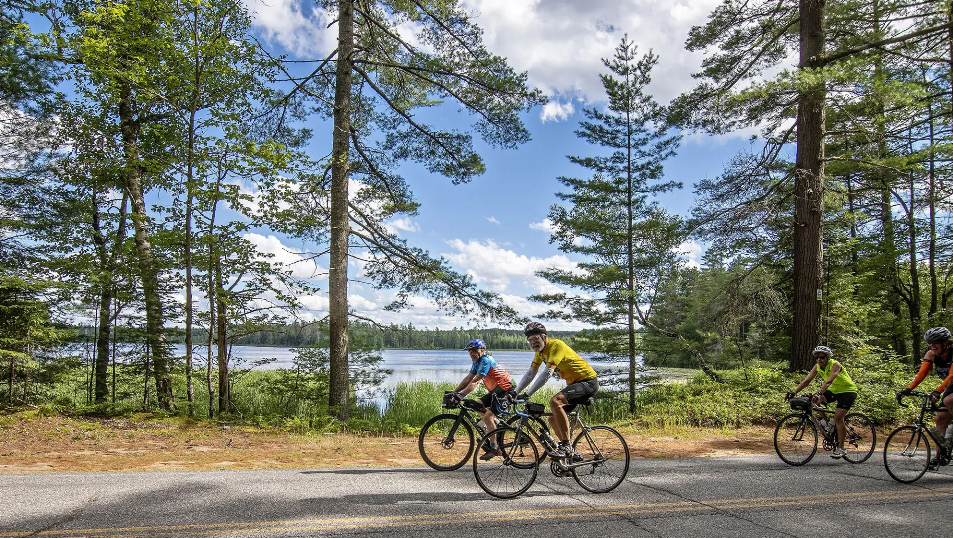 Cyclists wearing helmets and racing clothes ride on a flat path amid pine trees on a lakeshore.