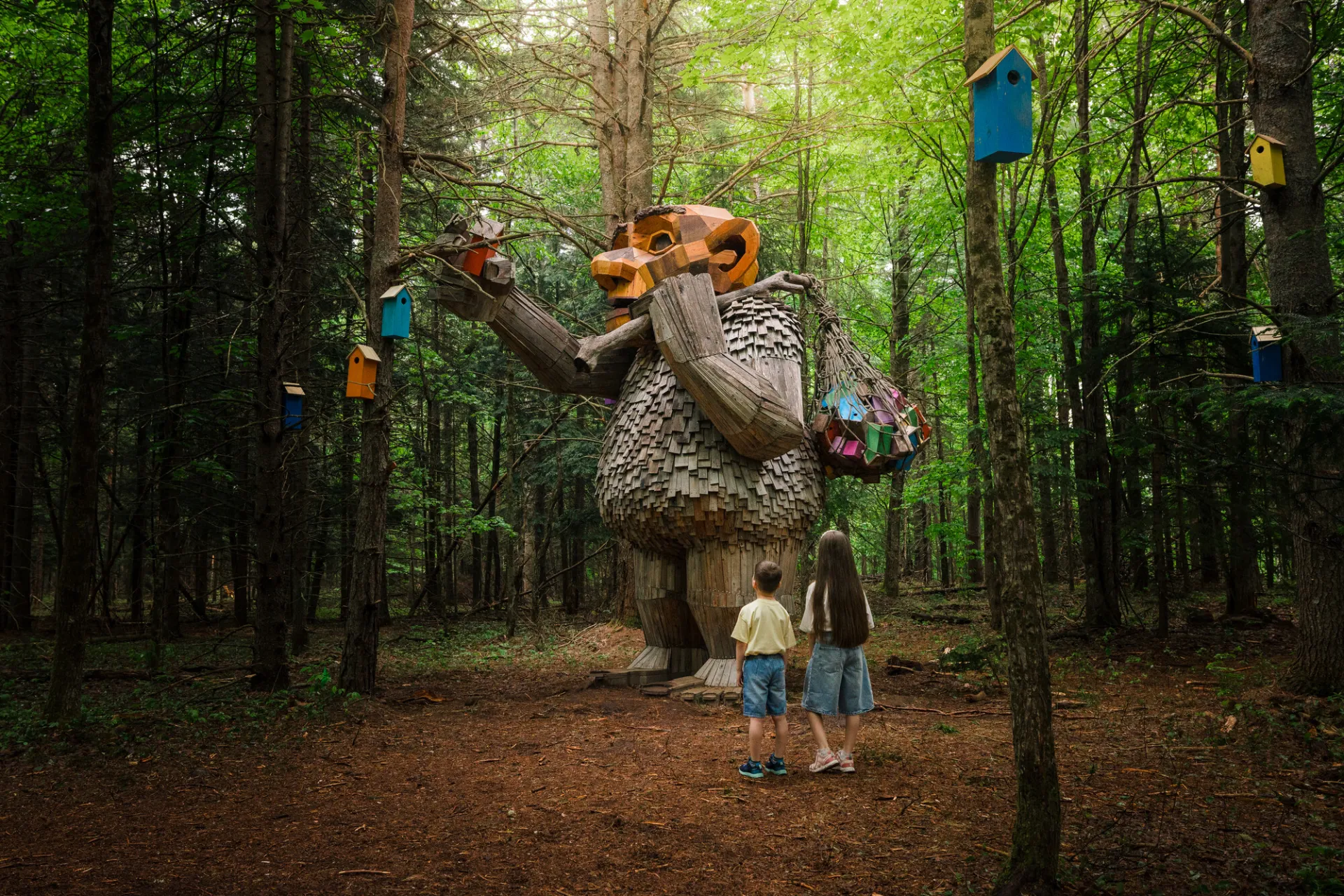 Two young children stand in the middle of the woods looking up at a wooden statue of a