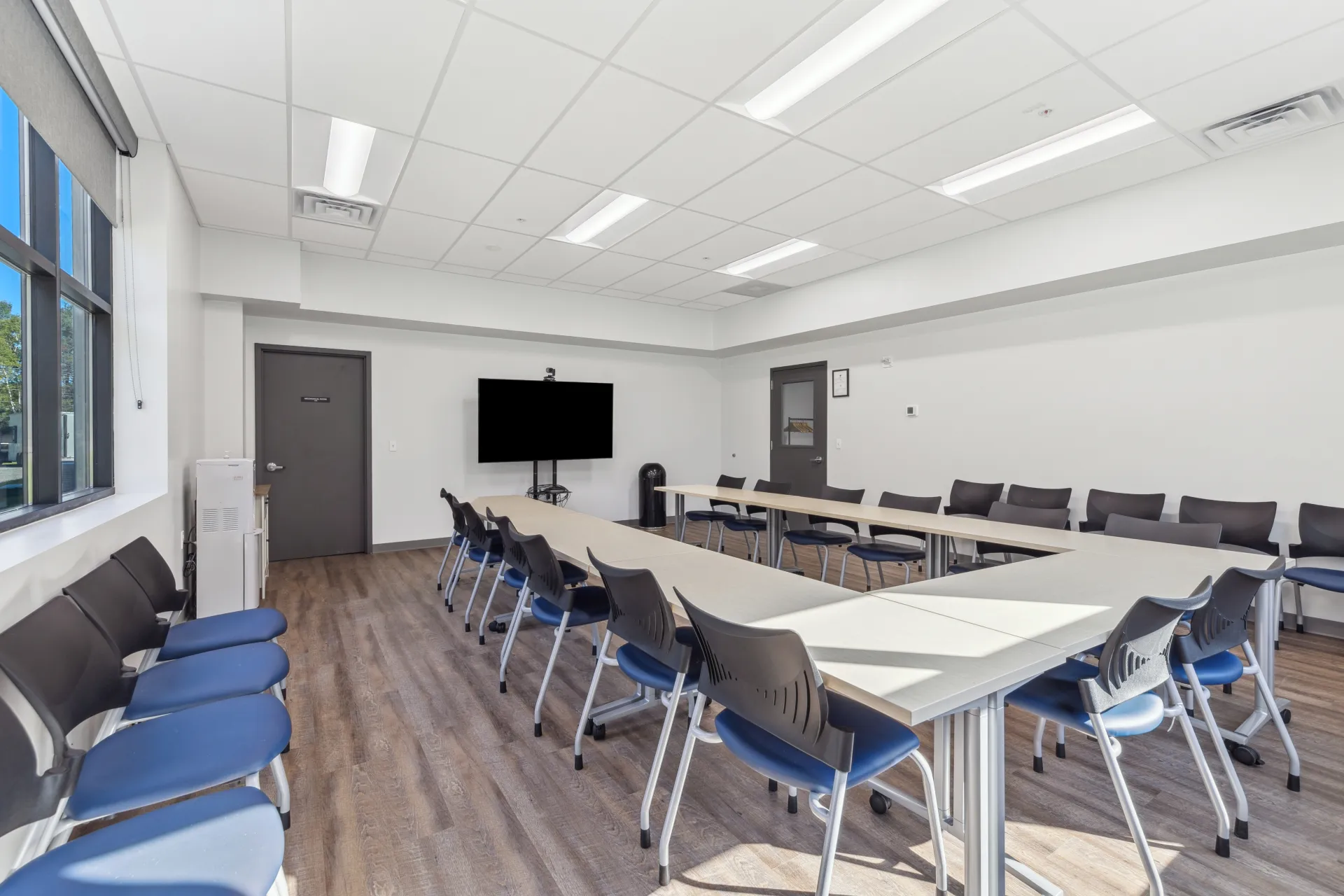 The meeting space arranged in a U shape with chairs and a large screen on a mount, viewed from the corner opposite the door.