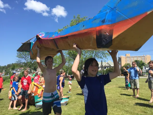 two boys holding a cardboard boat over their head as other look on in the background 