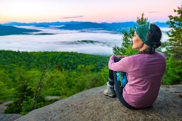 Woman in a purple shirt sits on the edge of Baker Mountain and stares out overtop of the green trees below with light cloud cover and other Adirondack mountains in the distance