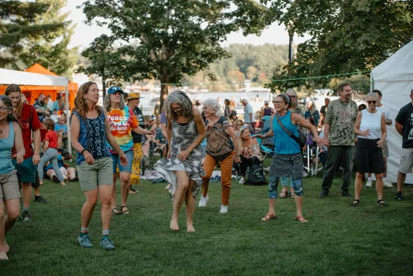A group of people dancing in the grass surrounded by families and vendor tents on the green by Lake Flower in Saranac Lake.