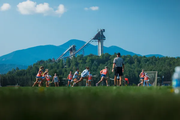 Two teams of women play lacrosse on field in front of mountains and ski jumps while a referee looks on