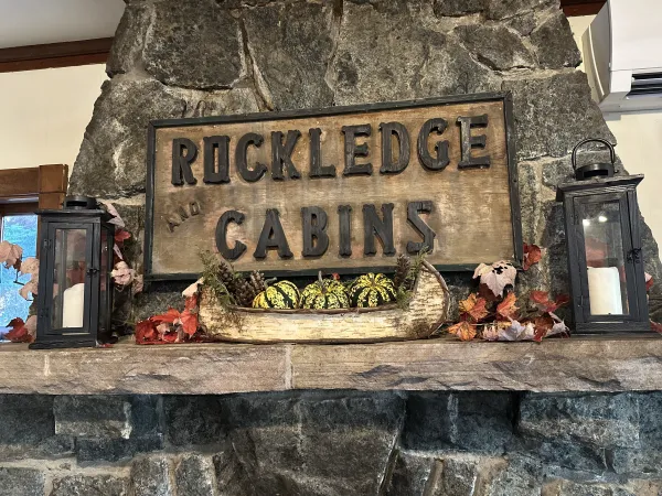 A carved sign reading 'Rockledge and Cabins' over a stone mantle with birch bark boat filled with festive squash and candles.