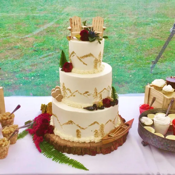 A three tiered cake frosted with an abstract mountain design and topped with adirondack chairs.