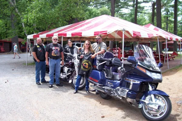 A group of motorcycles and their riders in front of the tent and picnic tables.