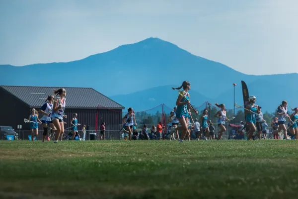 Female lacrosse players from two teams run down field with Whiteface Mountain in background