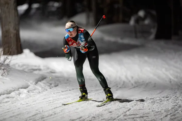 A cross-country skier in a racing suit and sunglasses glides along a snowy trail at night under bright artificial lighting.