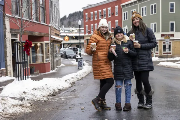 Three women standing in the middle of a winter village smile with hot cocoa in their hands. 