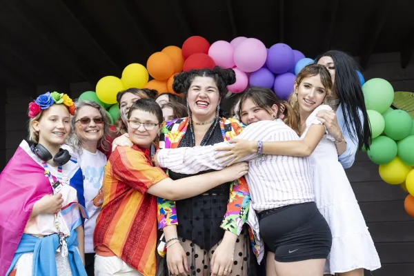 A variety of people hugging in front of a rainbow arch of balloons.