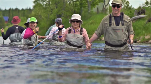 A group of people in chest wadders and walking sticks journey through a river