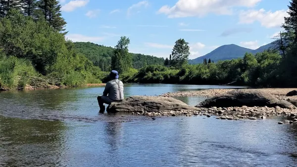A man sits on a rocky outcropping with his feet in the cool river