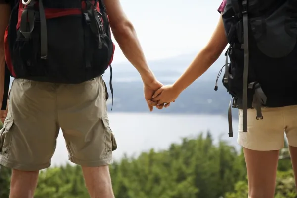 Two people hold  hands with backpacks on and looking at a view with trees and a body of water.