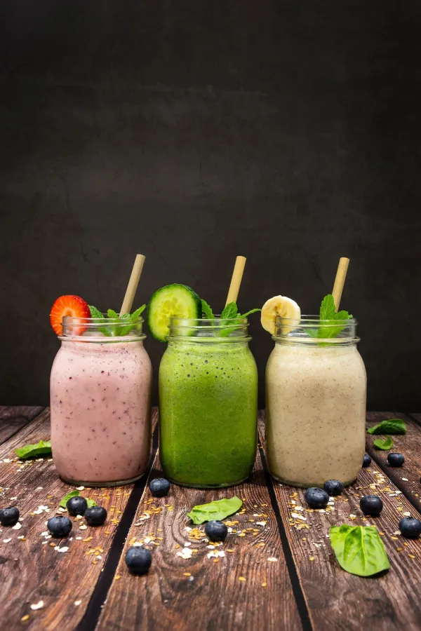 Three colorful mason jars of fresh cold pressed juice on a table surrounded by mint leaves and blueberries.