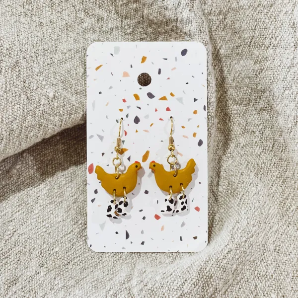A pair of dangling earrings featuring a golden chicken wearing cow print boots.