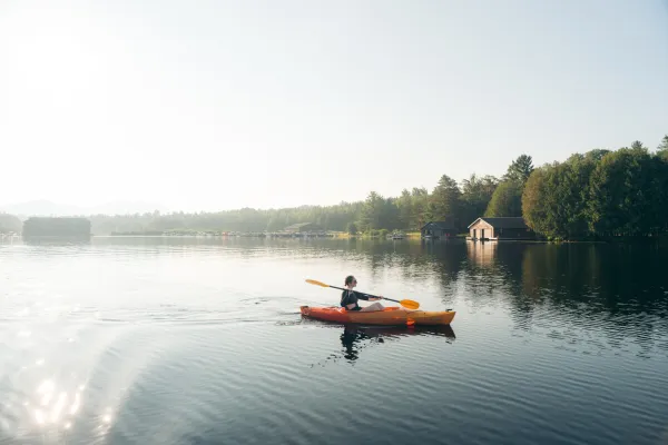 Woman in long sleeve rash guard and sunglasses paddles red and orange kayak on placid lake with shoreline house in background