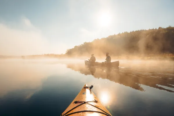 Two people paddle canoe on mirror-like water with fog seen past bow of orange kayak