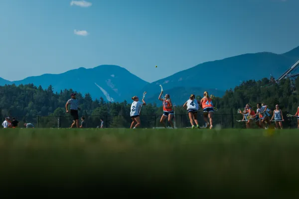 Two female lacrosse players look up at yellow lacrosse ball during play on field in front of Adirondack Mountain backdrop