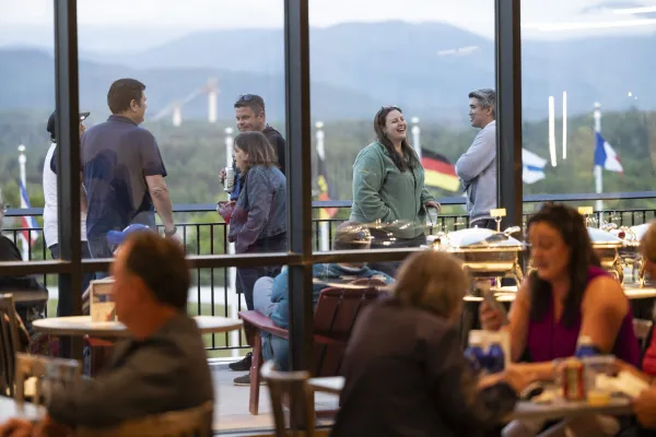 People seen mingling inside and outside of big glass windows with view of Lake Placid from outside deck