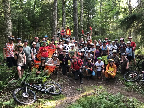 Large group of people, many dressed in a combo of animal costumes and biking gear, pose for photo in the woods with bikes