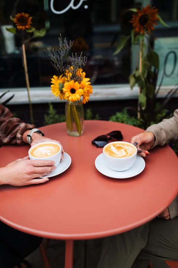 Two lattes sit on a colorful table with hands extending to retrieve them.
