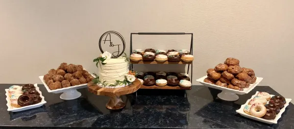 A single tier cake with an 'A & J' topper, flanked with trays of donuts.