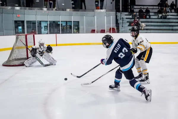 An ice hockey player in a navy blue jersey takes a shot on goal while a defender and goalie prepare to block the puck.