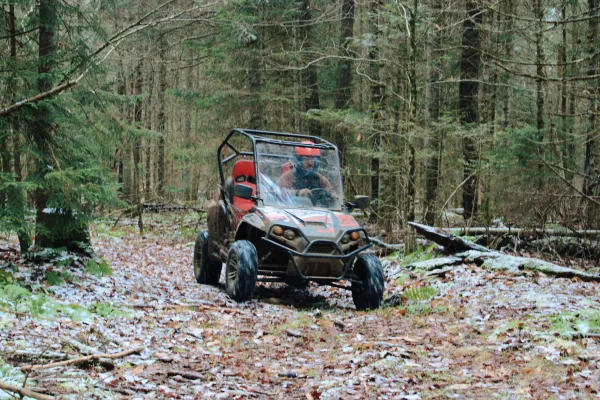Early snow covers the fallen leaves as a man in a helmet drives a UTV on a path through the trees 