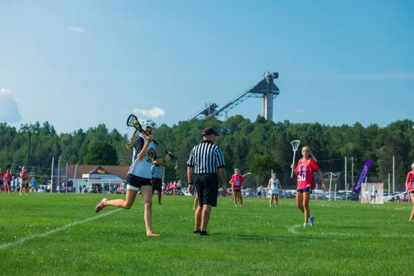 Woman in white and black uniform holds lacrosse ball in stick while running past referee during game on field in Lake Placid