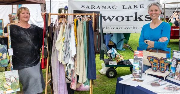 Two women stand by a table and a display rack of clothing outside under a tent with a banner reading Saranac Lake artworks behind them