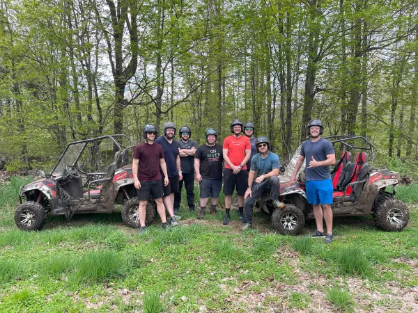 Eight happy young men stand together wearing helmets and smiling between all terrain vehicles in a grassy patch