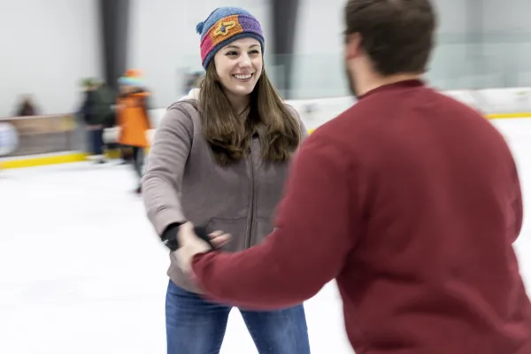 A smiling couple holding hands tentatively on the ice.