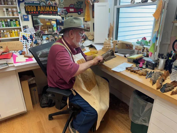 An older gentleman sits at a desk wearing a felted cap and a leather apron while examining a piece of raw wood