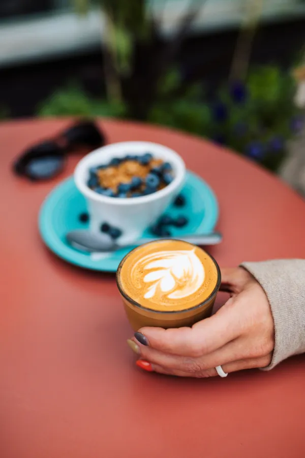 A hand holds a latte on a table next to a breakfast of berries and granola over yogurt.