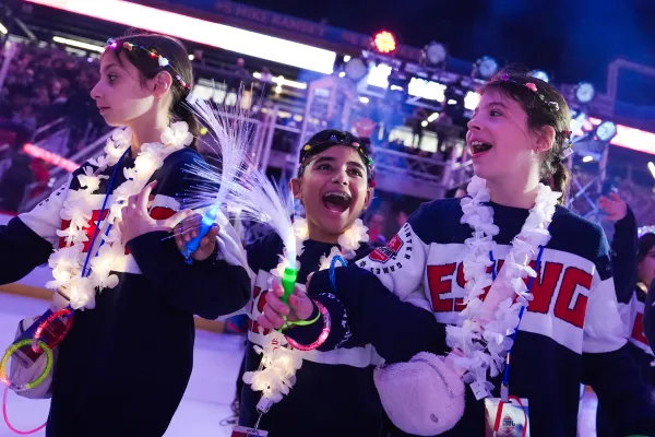 Three smiling children wearing "ESWG" sweaters celebrate with light-up wands during an opening ceremony.