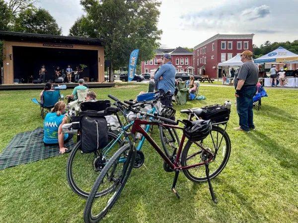 A group of adults with bicycles and picnic blankets gather on the green lawn of a village park.
