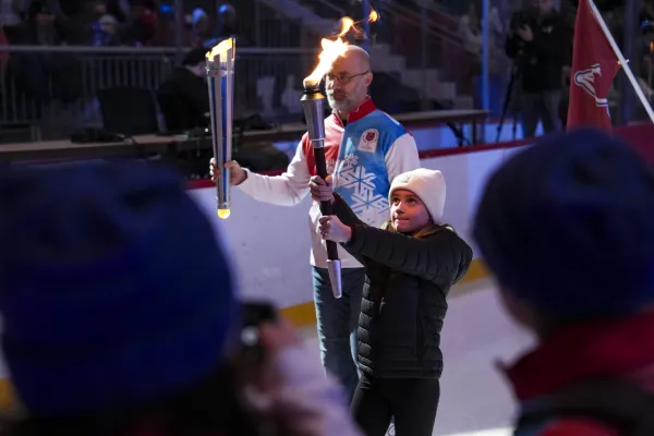 A young girl in a white beanie holds a flaming torch, assisted by an older man in a blue and red jacket. 